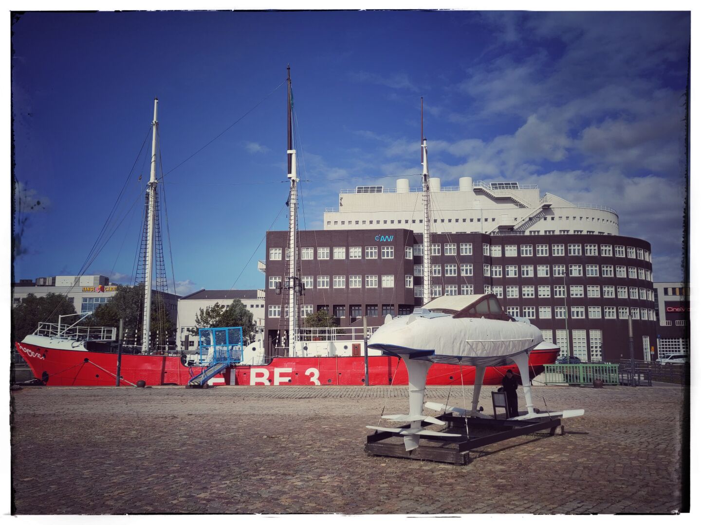 A red historic ship with three tall masts is moored in Bremerhaven’s Old Harbour. A small white vessel or submarine model stands on the cobblestone square in the foreground. Behind the ship is the large brown-and-white building of the Alfred Wegener Institute beneath a deep blue sky with scattered clouds.