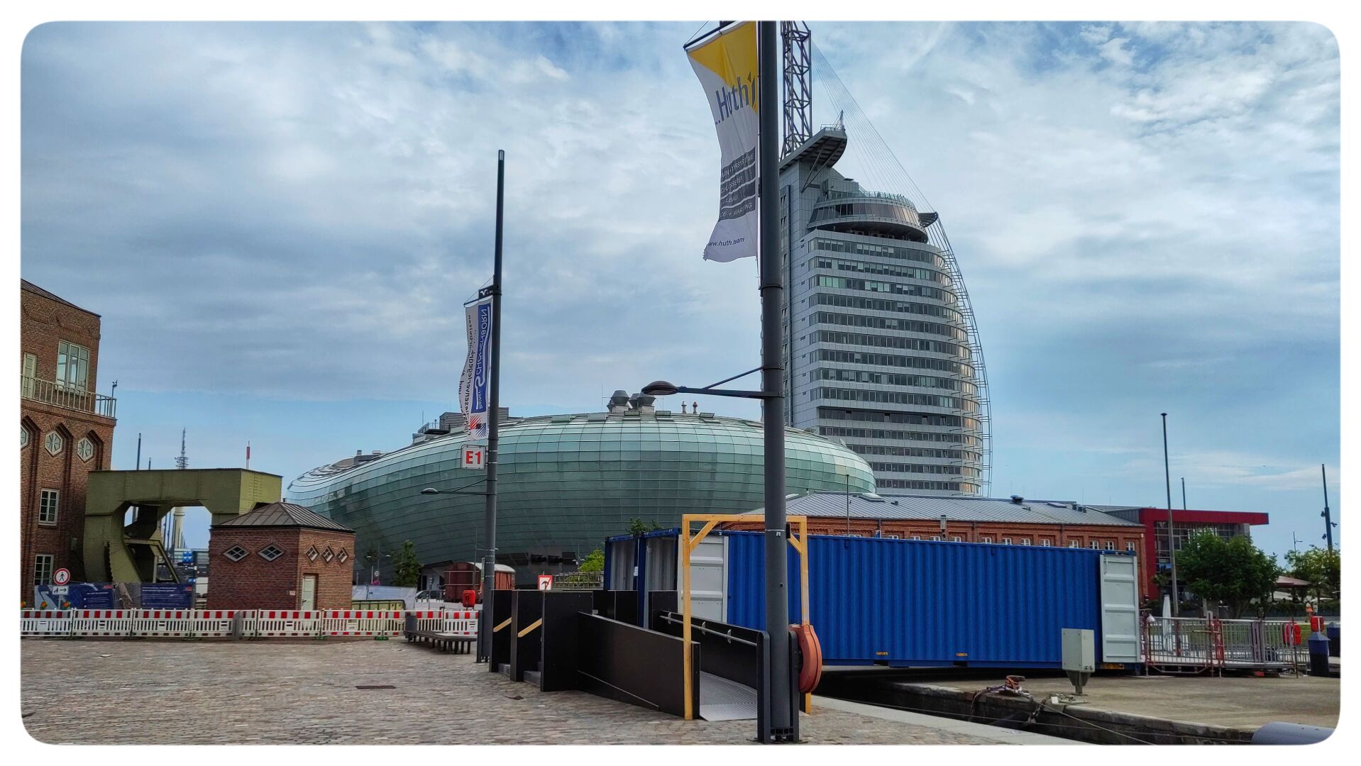 View of Bremerhaven’s Havenwelten under a cloudy blue sky. A temporary crossing with black ramps and a large blue structure spans the narrow canal in the foreground. Behind it are the curved green façade of the Klimahaus and the tall, sail-shaped Atlantic Hotel Sail City.