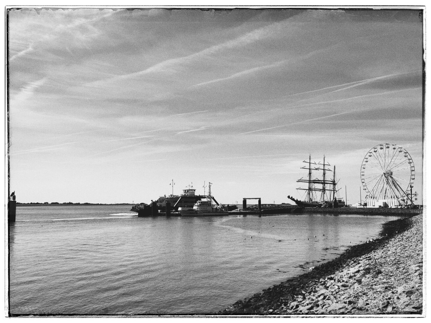 Black-and-white photo looking across the water from the Havenwelten towards Willy-Brandt-Platz in Bremerhaven during the Maritime Tage. Several harbor vessels and a large sailing ship are moored near the quay, with a Ferris wheel rising prominently on the right. A wide sky with long streaks of clouds dominates the background.
